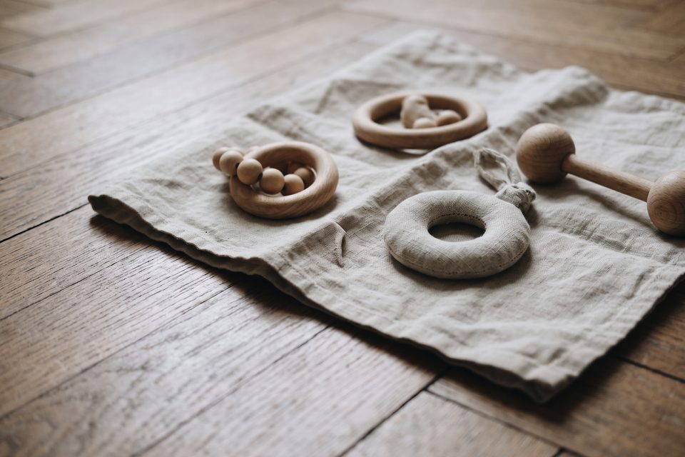 Deux hochets en bois brut et un hochet en tissu uni posés sur un lange de lin, parquet de chêne flou au premier plan