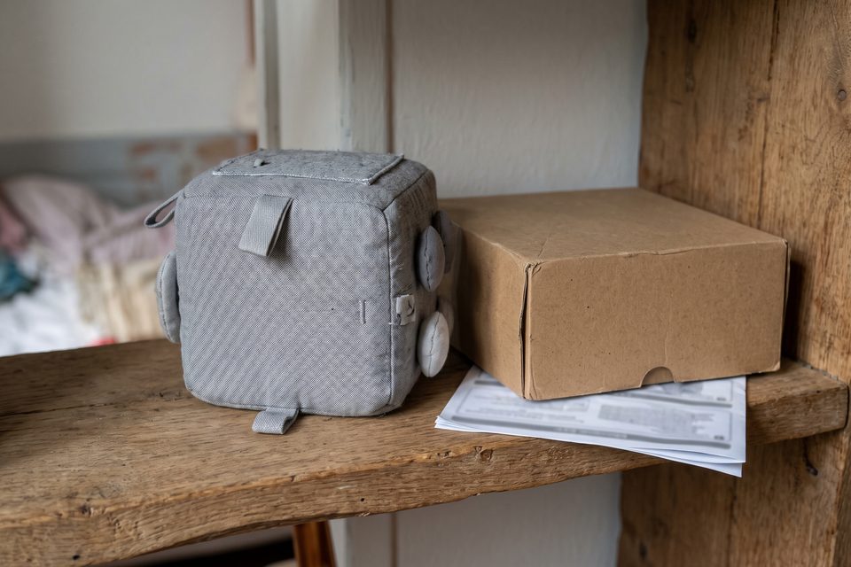 Cube d'éveil en tissu uni posé à côté de son emballage en carton retourné, sans texte lisible, sur une étagère basse en bois