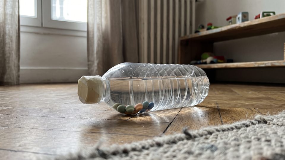 Bouteille transparente remplie d'eau et de perles colorées, posée sur un parquet près d'une fenêtre