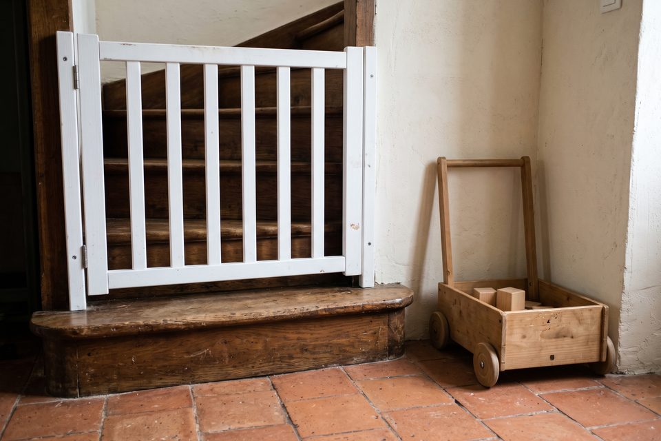 Bas d'un escalier en bois avec une barrière de sécurité fermée, un chariot de marche en bois rangé contre le mur, tomettes au sol