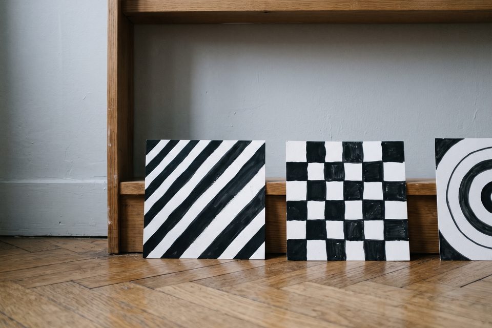 Trois cartes à motifs noir et blanc appuyées contre une étagère basse en bois, au ras d'un parquet de chêne, lumière grise d'une fenêtre